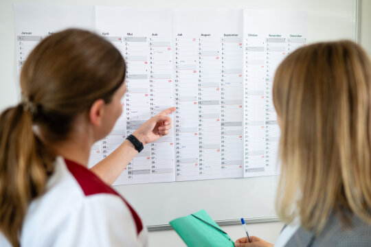 Eine Hauswirtschafterin in Arbeitskleidung und eine Hauswirtschafterin in Blazer stehen vor einem Wandkalender.