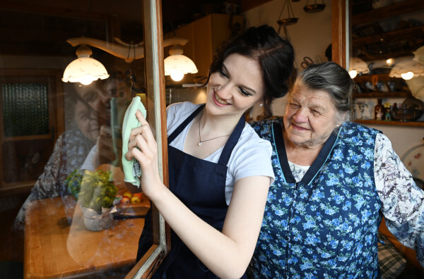 Junge Hauswirtschafterin reinigt ein Fenster bei einer Seniorin.