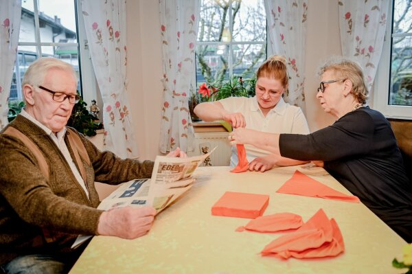 Seniorin faltet mit Pflegerin Servietten an einem Esstisch, Senior sitzt dabei und liest Zeitung.