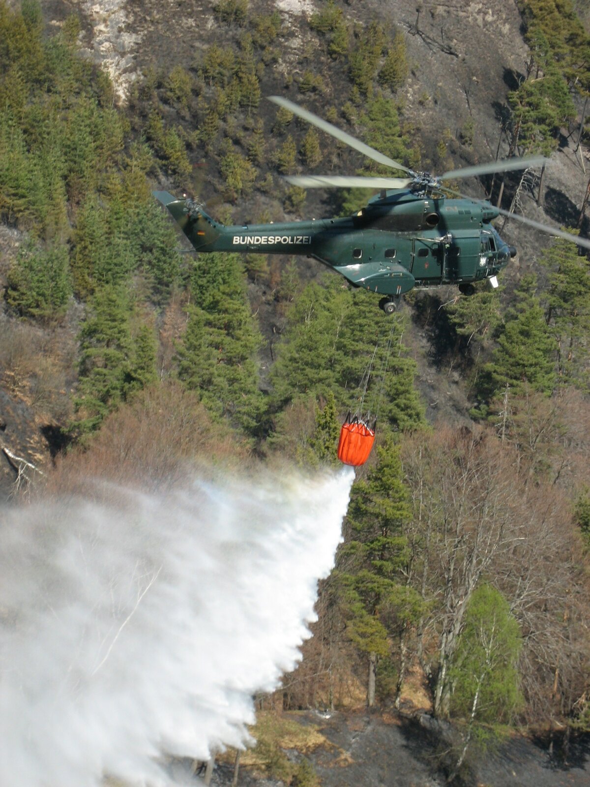 Das Bild zeigt einen Löschhubschrauber im Flug über einem brennenden Wald, er wirft Wasser ab.