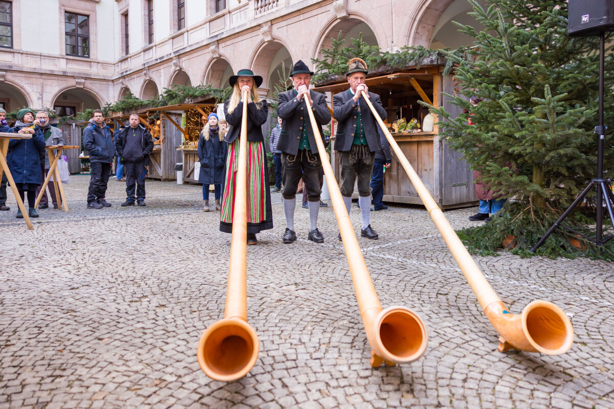 Zwei Alphornbläser und eine Alphornbläserin im Innenhof des Ministeriums mit den Ständen zum Hoffest im Advent.