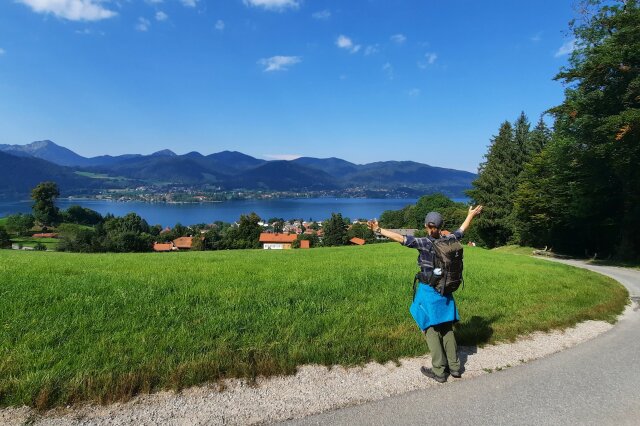 Eine Wanderin auf einem Fußweg, im Hintergrund sieht man eine Landschaft mit Dörfern, Seen und Gebirge.