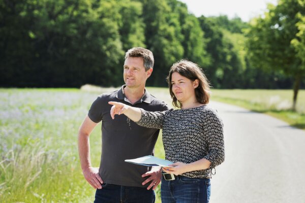 Ein Mann und eine Frau stehen draußen auf einem landwirtschaftlichen Weg. Die Frau hält ein Dokument in der Hand und zeigt mit dem Finger in die Ferne.