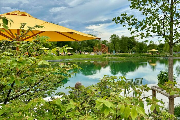 Büsche im Vordergrund mit gelben Sonnenschirm und See im Hintergrund. Aufgenommen auf der Gartenschau in Heimstetten.