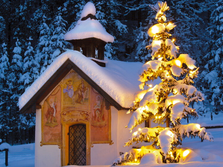 Verschneite Kapelle mit einem beleuchteten Christbaum in der Abenddämmerung.