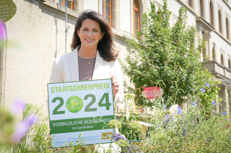 Staatsministerin Michaela Kaniber im Klimagarten des Ministeriums mit einem Schild zum Staatsehrenpreis vorbildliche Ausbildung Gartenbau 2024.