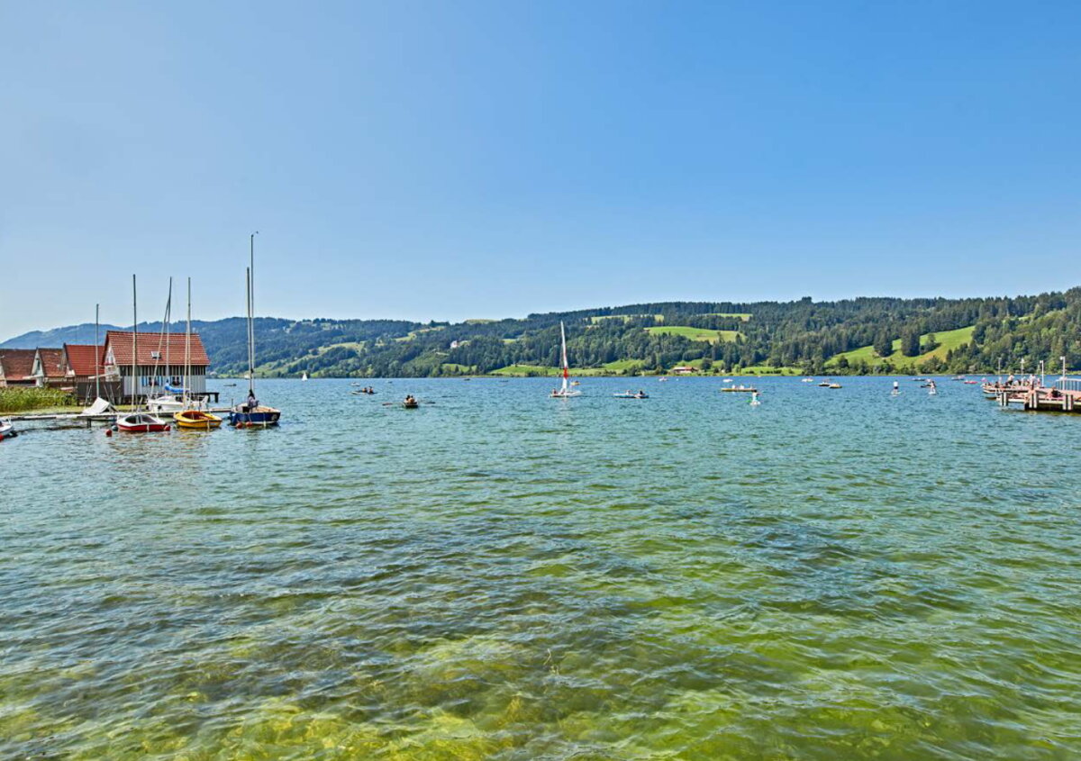 Bayerischer See mit Segelbooten und Steg. Im Hintergrund Hügel mit Bäumen.