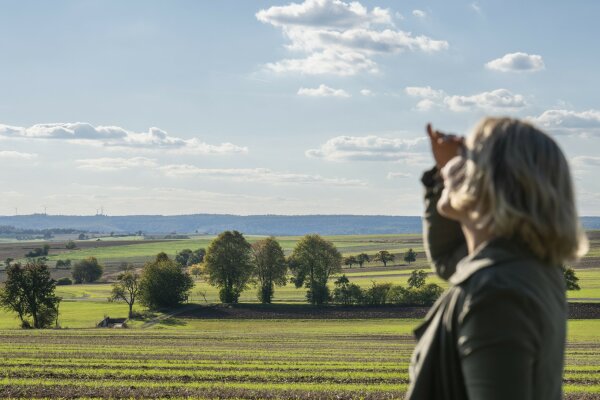 Frau vor schöner Landschaft hebt die Hand über die Augen um in die Ferne zu sehen.