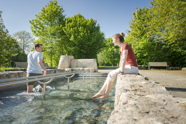 Junge Mann steht in einem niedrigen Beck und watet durch das Wasser. Eine junge Frau sitzt am Beckenrand