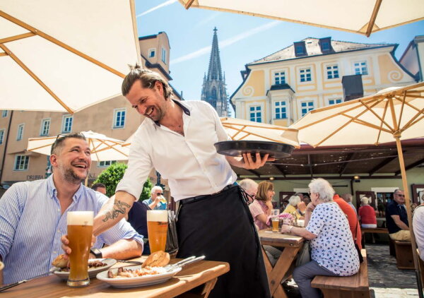 Ein Mann sitzt fröhlich in einem Biergarten. Auf dem Tisch steht das Essen. Der Kellner bringt ein Bier. 